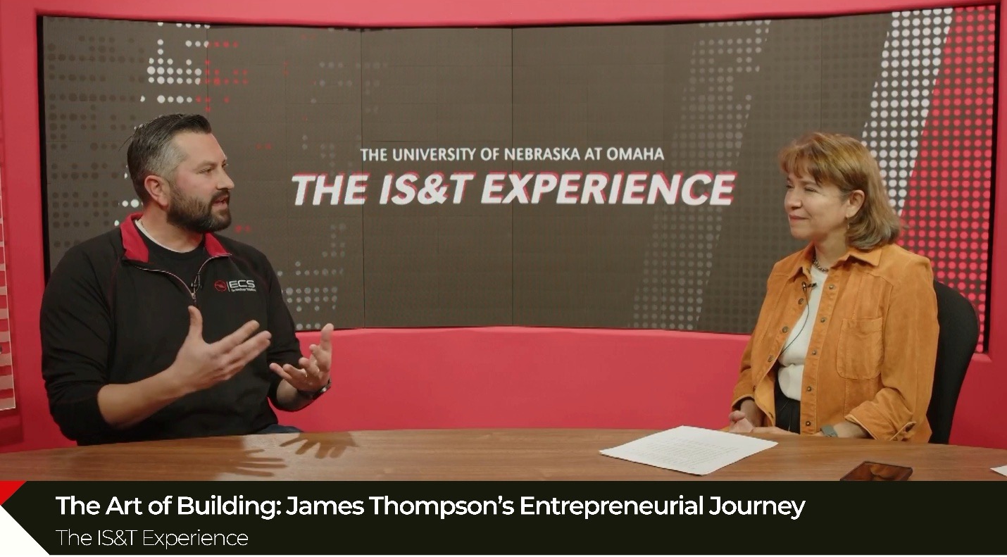 Photograph of a studio interview featuring two people seated at a table with a large screen behind them displaying "The University of Nebraska at Omaha THE IS&T EXPERIENCE," with a caption at the bottom reading "The Art of Building: James Thompson’s Entrepreneurial Journey" and "The IS&T Experience."