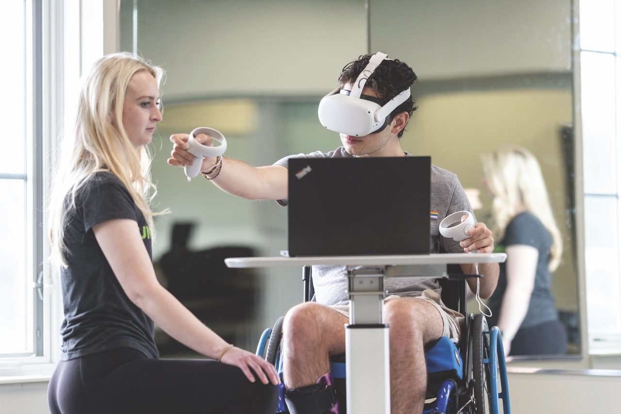 A wheelchair user tests a VR headset and controllers while a researcher observes at a workstation.