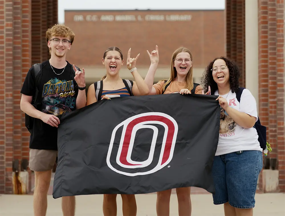  Four students stand outside the Criss Library holding a large black flag with a red and white “O” logo, showing school pride on the University of Nebraska Omaha campus.