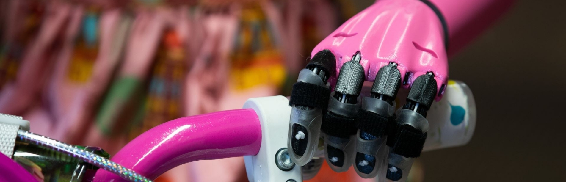 This image shows a close-up of a prosthetic hand attached to the handlebars of a bicycle. The hand has a pink color theme and is equipped with black straps designed to grip the handlebar securely. The bicycle itself seems to have a pink grip and a patterned bell, suggesting a playful or child-friendly design. The focus on the mechanical hand and its integration with the bicycle highlights an assistive technology for bike riding.