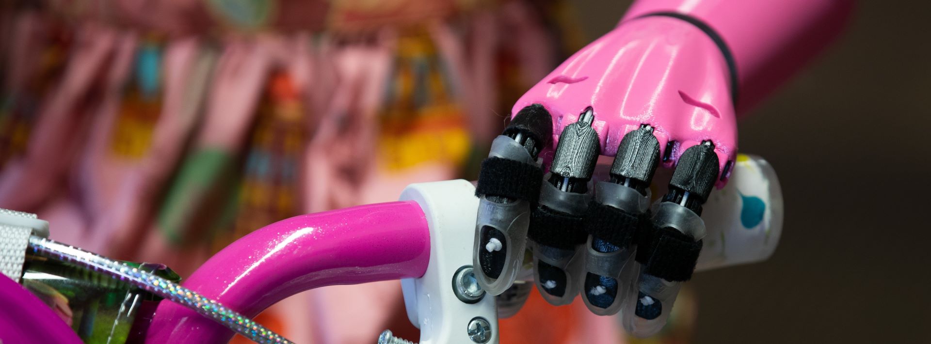 This image shows a close-up of a prosthetic hand attached to the handlebars of a bicycle. The hand has a pink color theme and is equipped with black straps designed to grip the handlebar securely. The bicycle itself seems to have a pink grip and a patterned bell, suggesting a playful or child-friendly design. The focus on the mechanical hand and its integration with the bicycle highlights an assistive technology for bike riding.