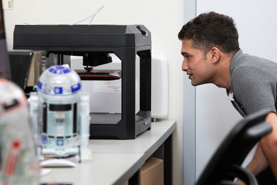  A person is intently observing a 3D printer at work in an office environment. Two models of astromech droids, resembling R2-D2, are in the foreground, slightly out of focus. The 3D printer is actively printing an object, indicated by the movement of its components. The person seems focused and curious, possibly monitoring the printing process or waiting for the completion of a project. The scene suggests a setting of innovation, technology, and design.