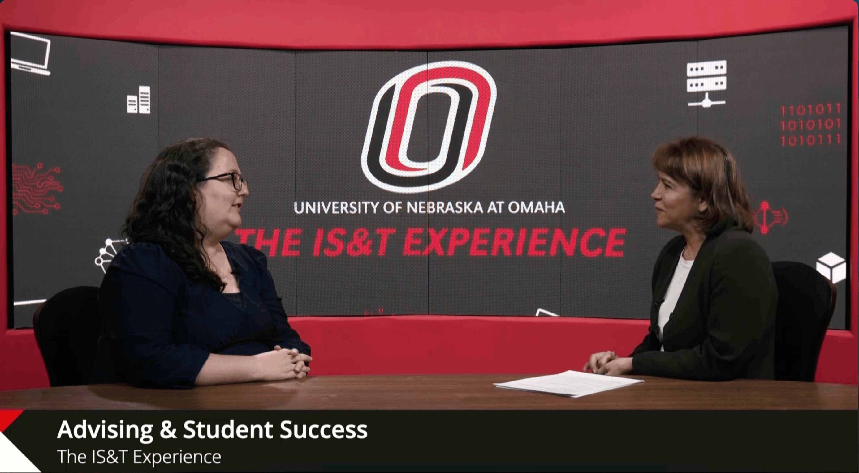 Two women in conversation at a table with the University of Nebraska at Omaha logo and 'THE IS&T EXPERIENCE' text behind them. Lower third reads 'Advising & Student Success.'