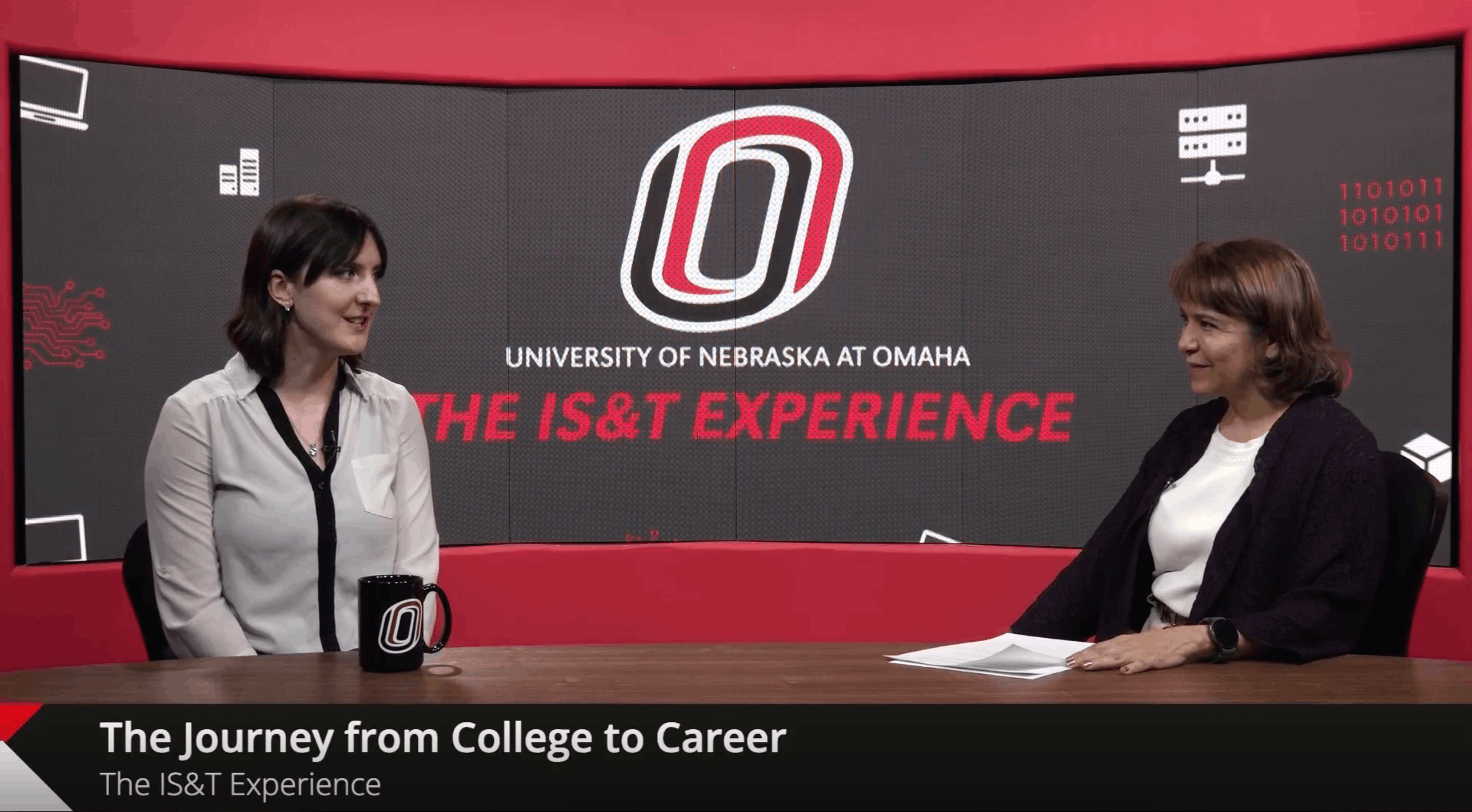 Two people in conversation at a table with the University of Nebraska at Omaha logo and 'THE IS&T EXPERIENCE' text behind them. Lower third reads 'Advising & Student Success.'