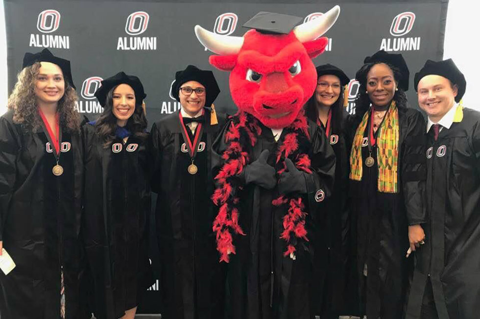 2018 doctoral program graduates in caps and gown, with Durango, the UNO mascot. 