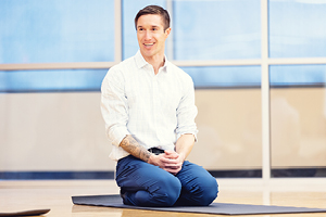 Man kneeling with his hands crossed on a mat in a gym.
