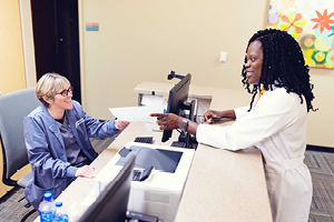 Healthcare office where one woman is sitting behind the front desk and another is handing her a piece of paper.