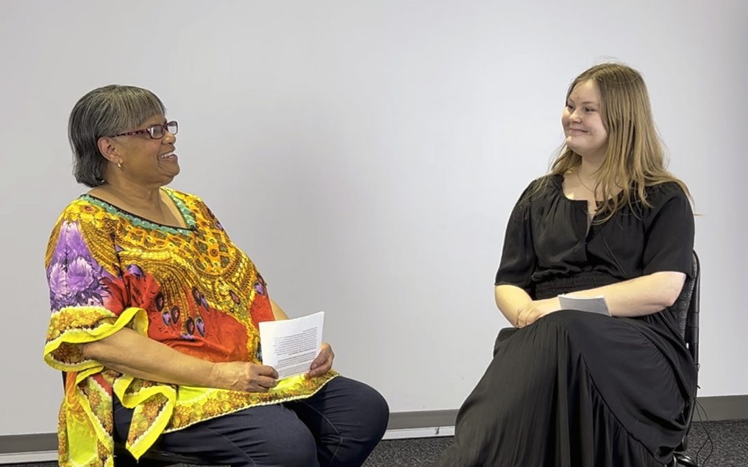 An older black woman talking to a younger white student with a white background behind them.