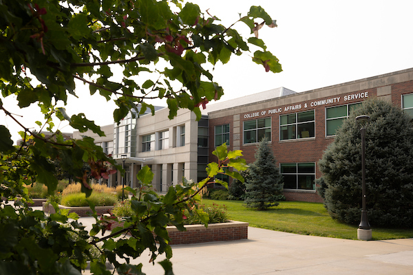 Exterior of the College of Public Affairs & Community Service building, framed by trees and landscaping on a sunny day.