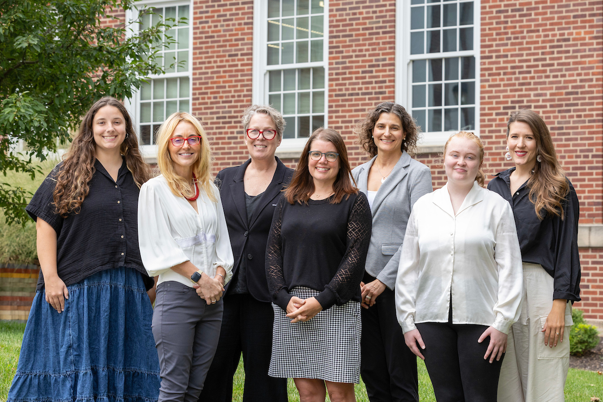 Seven women stand smiling in front of a brick building with large windows and greenery.