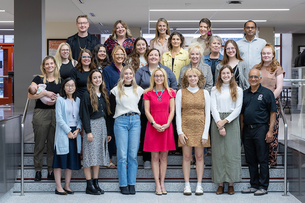 A large group of faculty, staff, and students stand smiling on indoor steps in a university building.