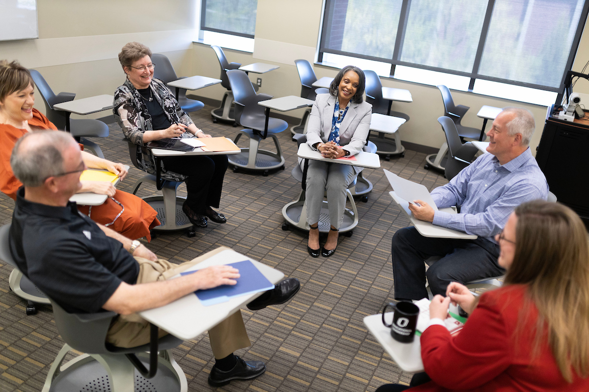 Six adults sitting in student desks in a circle.
