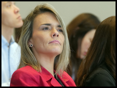 Woman in red jacket sitting in a classroom listening to a speaker.