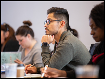 Man in gray sweater sitting at a desk with a group of people.