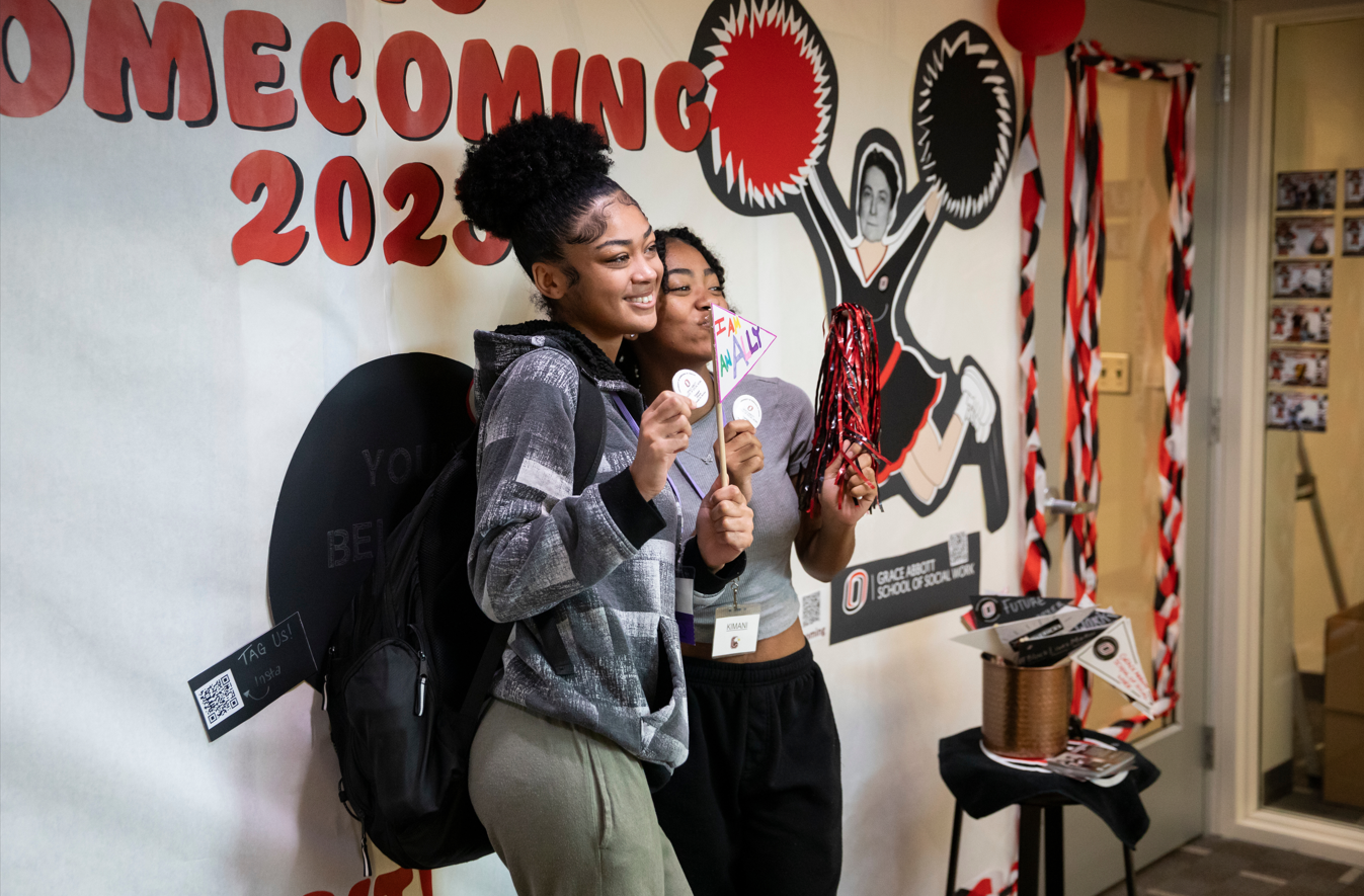 Two UNO students holding decorative flags in front of a homecoming poster.
