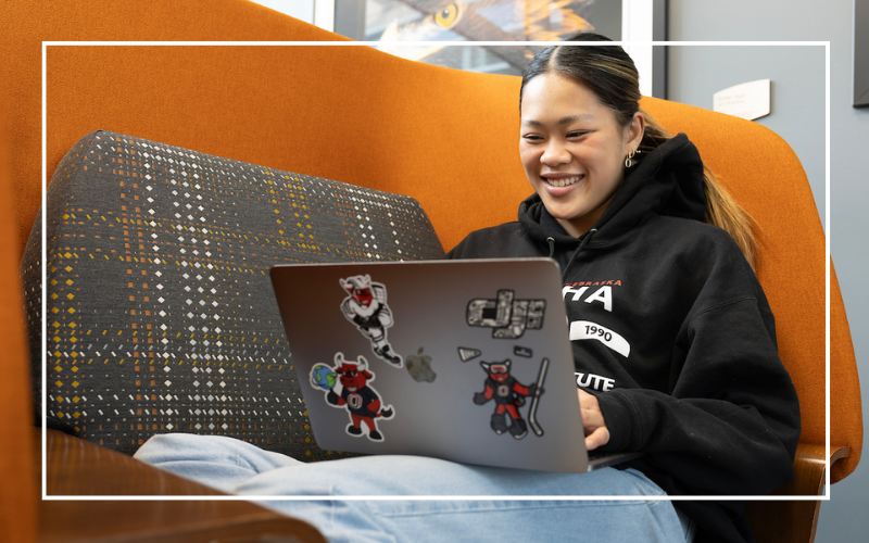 Student sitting on an orange lounge chair typing on a laptop.