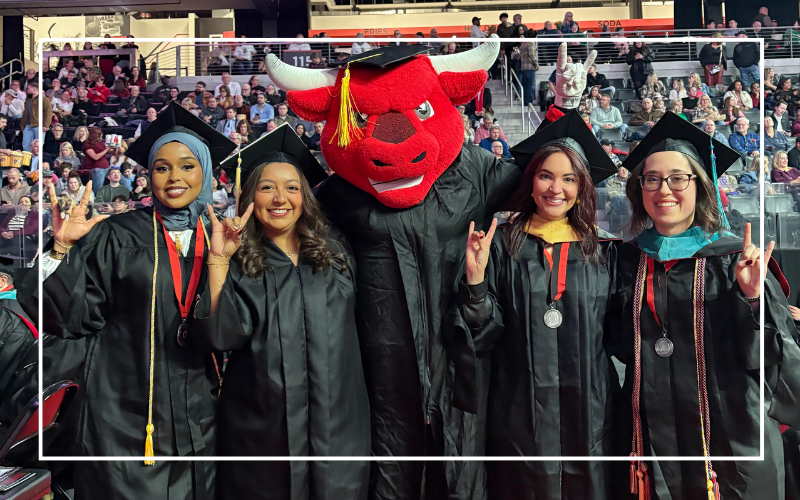 Four students at commencement posing with Durango in a cap and gown.