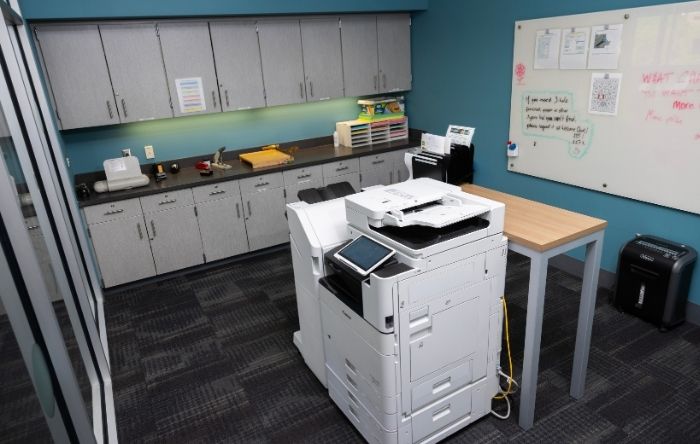 Shared printer room with a multifunction printer, storage cabinets, countertops, worktable, whiteboard, and paper shredder.