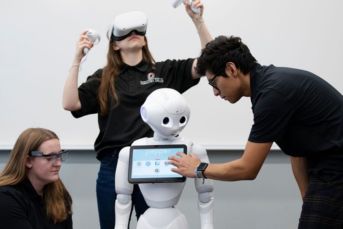 Three UNO students interact with a humanoid robot named Pepper using virtual and augmented reality tools during a research demonstration in the Koraleski Commerce and Applied Behavioral Lab.