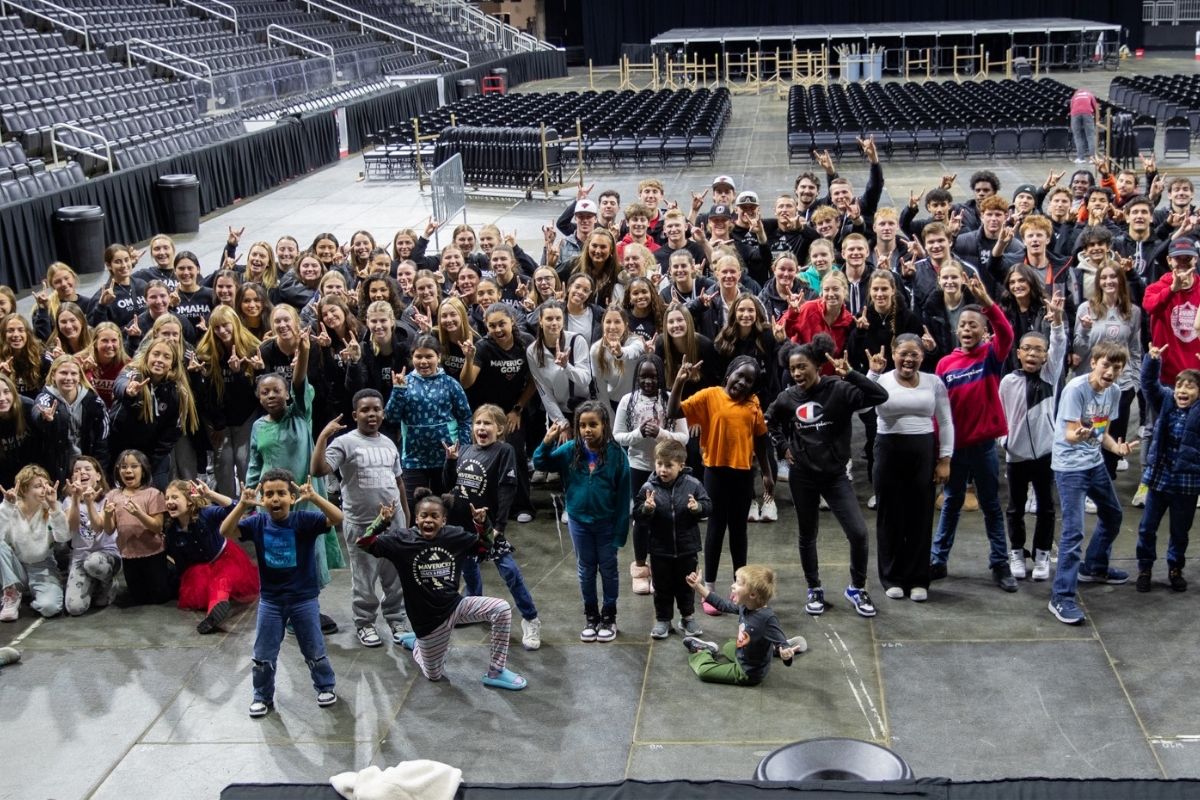 A large group of UNO student-athletes and youth gather on a gym floor, smiling and posing together after a community engagement event promoting teamwork and mentorship.