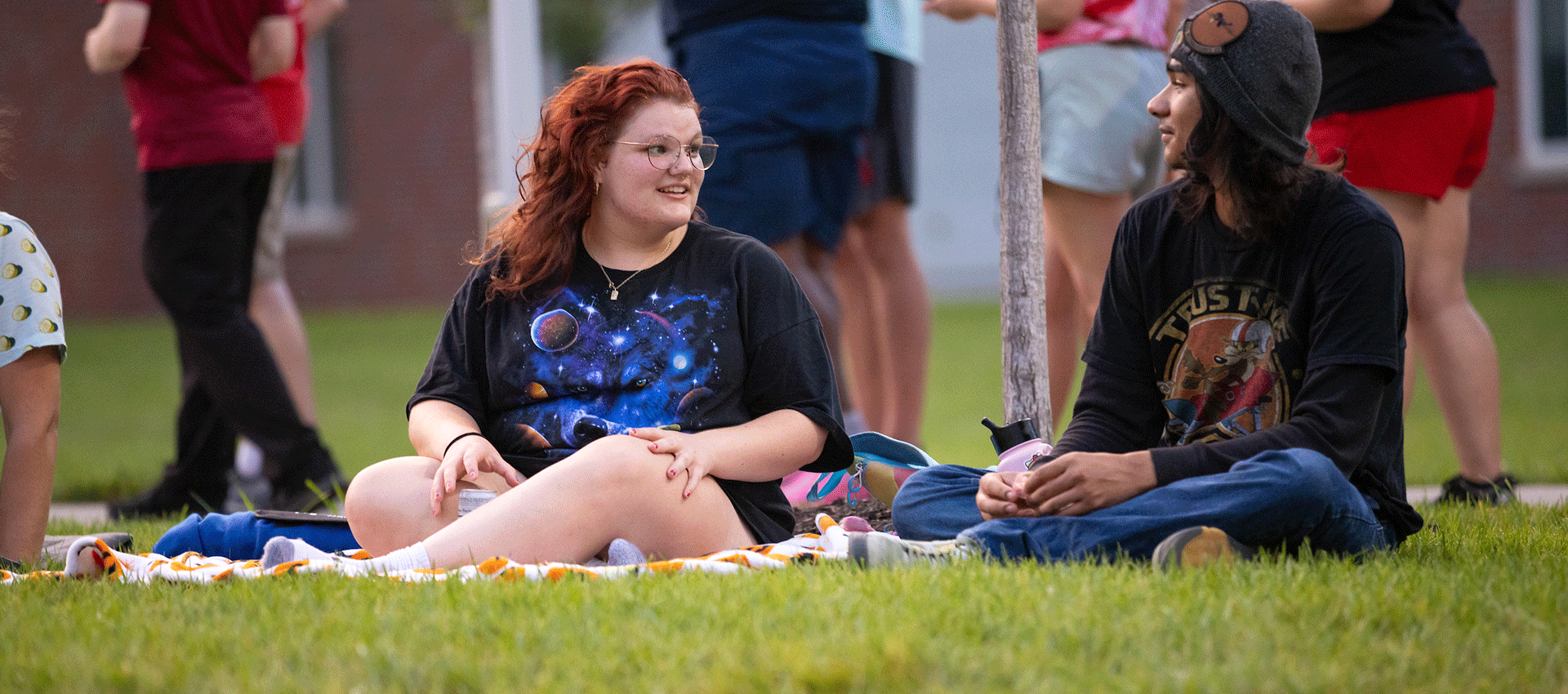 Two college students sit on a blanket on the lawn, talking during an outdoor campus event while others gather in the background.