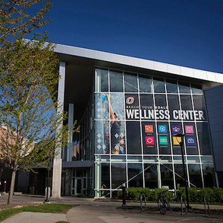 Exterior of the university’s Health and Wellness Center featuring a glass façade, large “Wellness Center” signage, and icons representing health and support services.