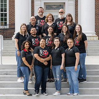 Group portrait of Counseling and Psychological Services staff standing together on the steps of a campus building, wearing matching CAPS shirts.