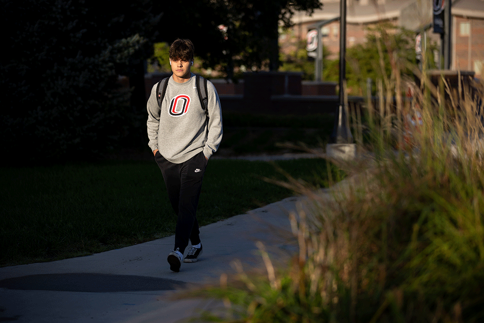 Student wearing a University of Nebraska Omaha sweatshirt and backpack walking along a campus sidewalk in warm evening light.