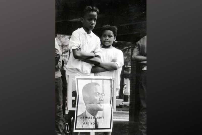 Two children with their arms crossed behind a sign bearing a photograph of Malcolm X with the text "He was Ready! Are you?"