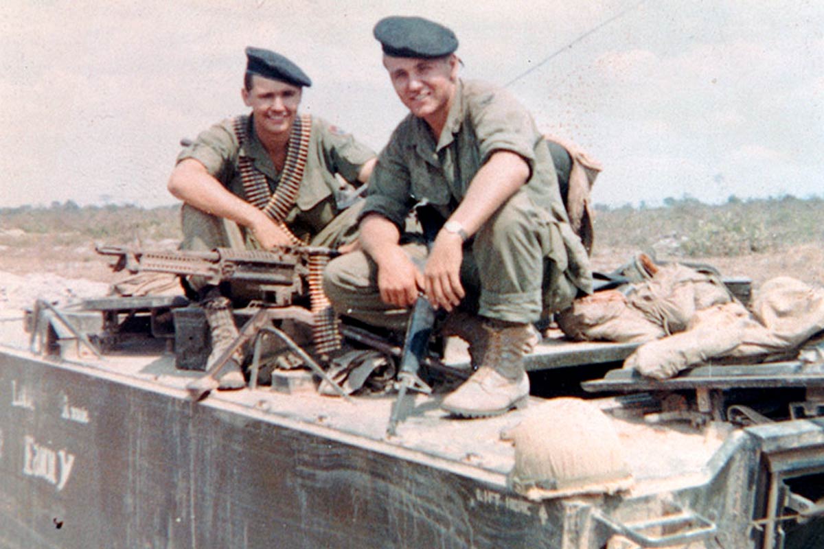 two men in army fatigues squatting on top of a military truck 