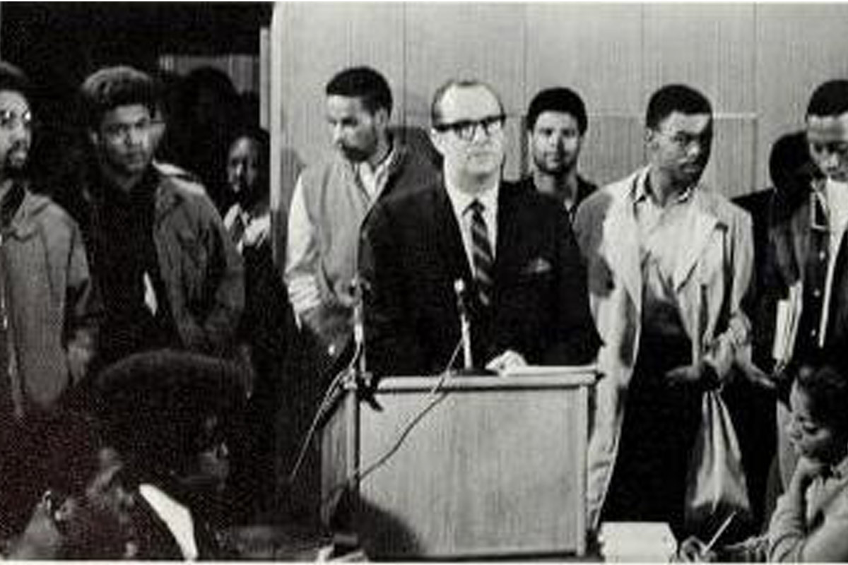 a man speaking at a podium with students surrounding him