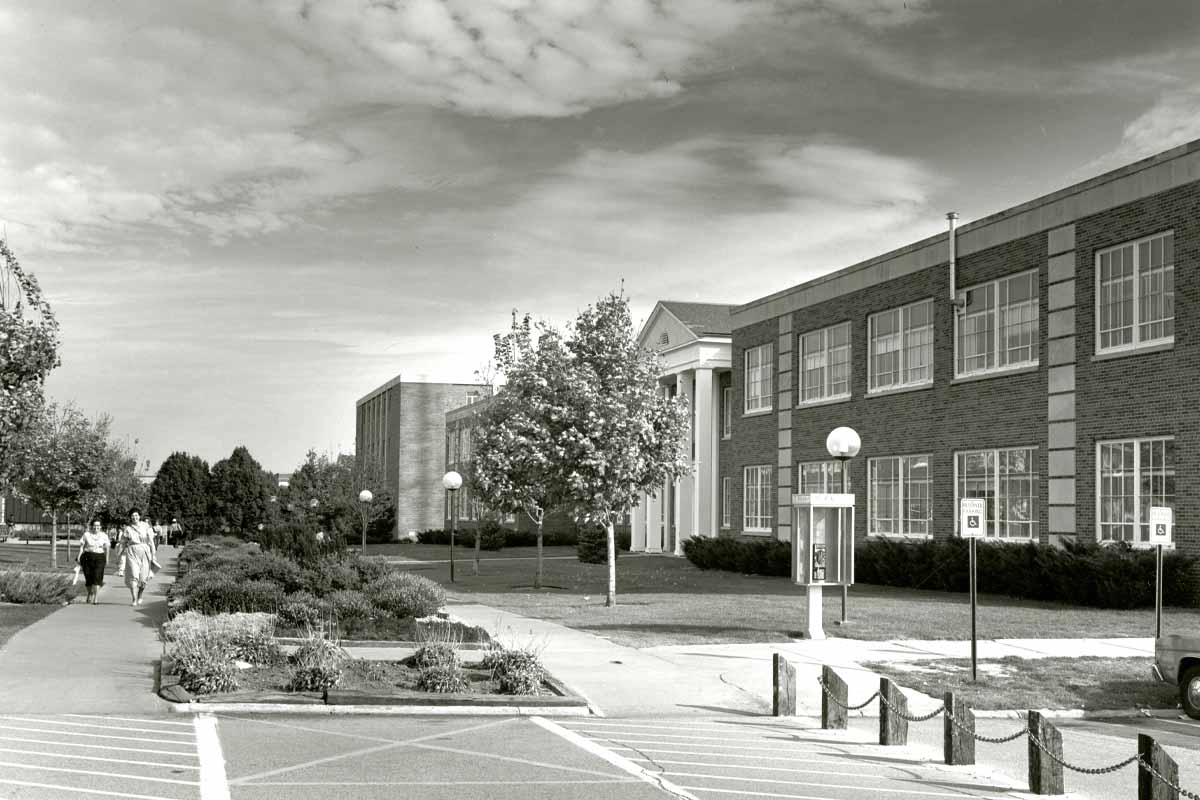 a black and white photograph with two women walking down the side on the left, and on the right the side of large brick building. There are two sidewalks with foliage in between them.