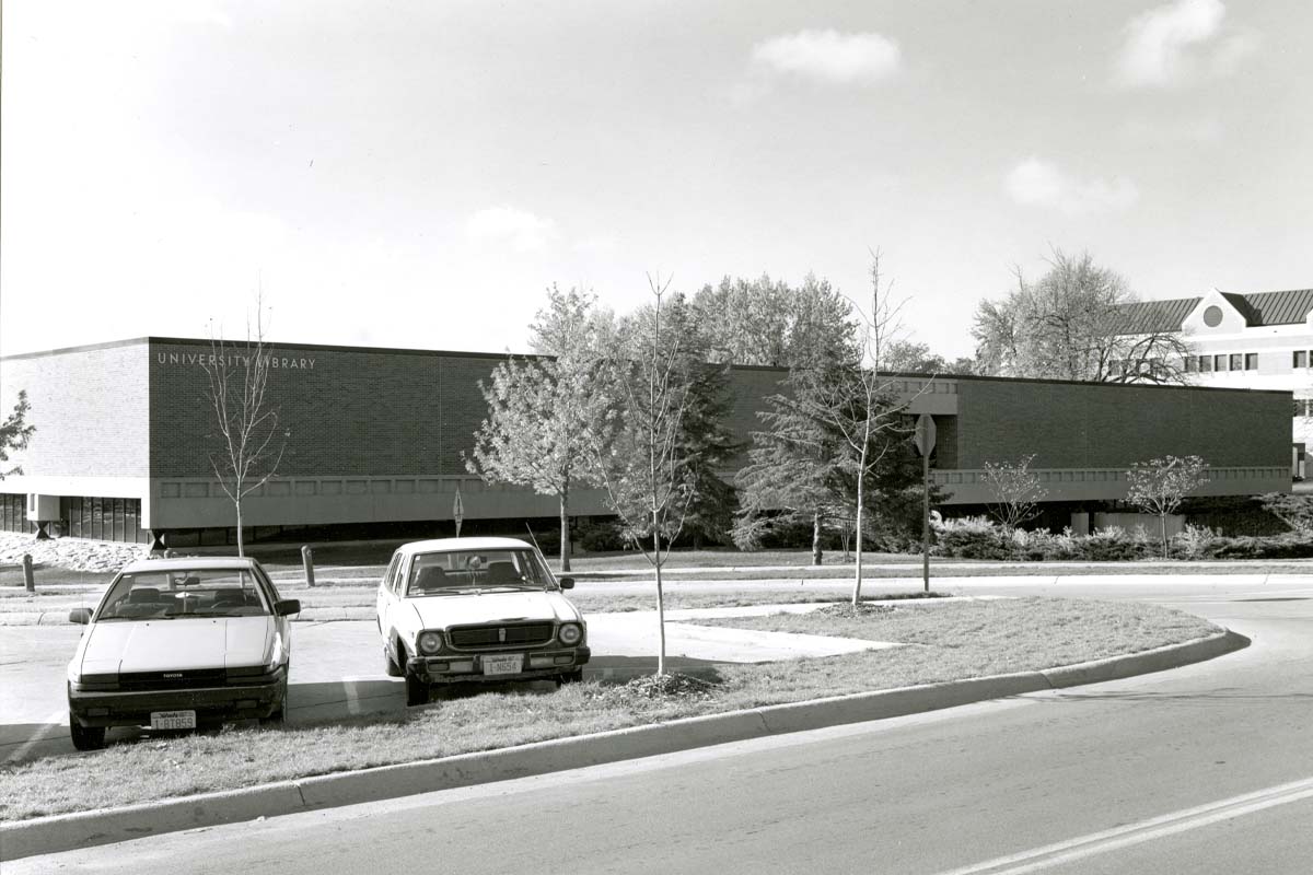 a black and white photograph of a brick building across the street with the words 'University Library' on the building, Two cars are parked in a parking lot near the building.