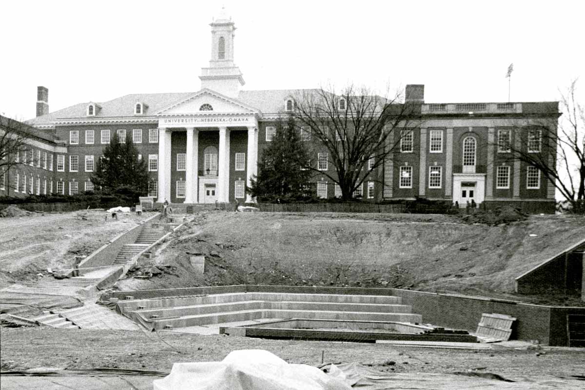 a black and white photograph of a large brick building with concrete stairs leading down to an open area. There are white columns on the front of the building and the words 'University of Nebraska at Omaha' is inscribed on the front of the building.