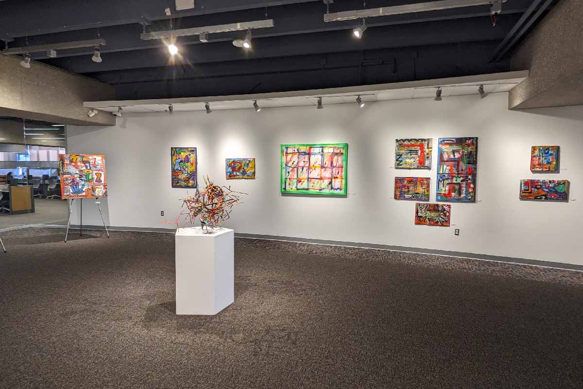 an art gallery with many colorful art canvases hanging on the wall. in the middle of the room there is a pedestal with a colorful sculpture on it.