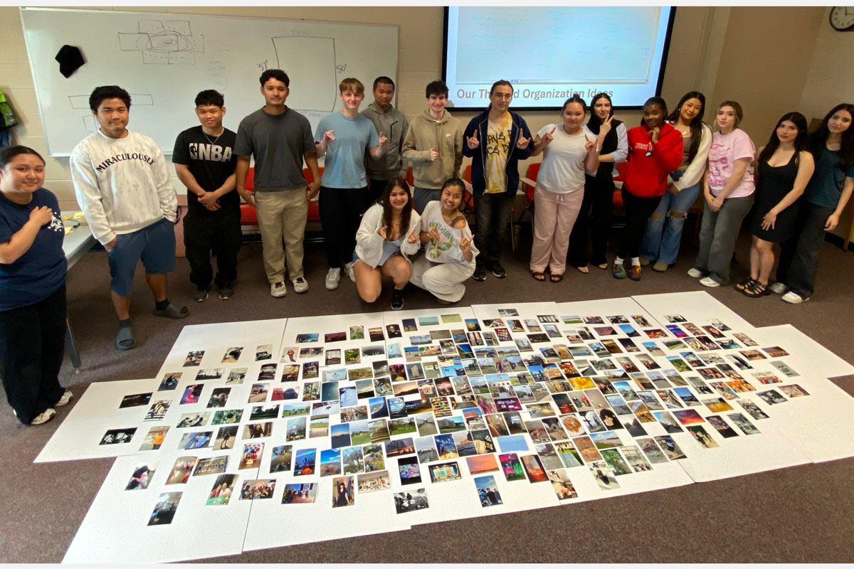 a group of college students standing around a photo collage showcasing their work and smiling at the camera