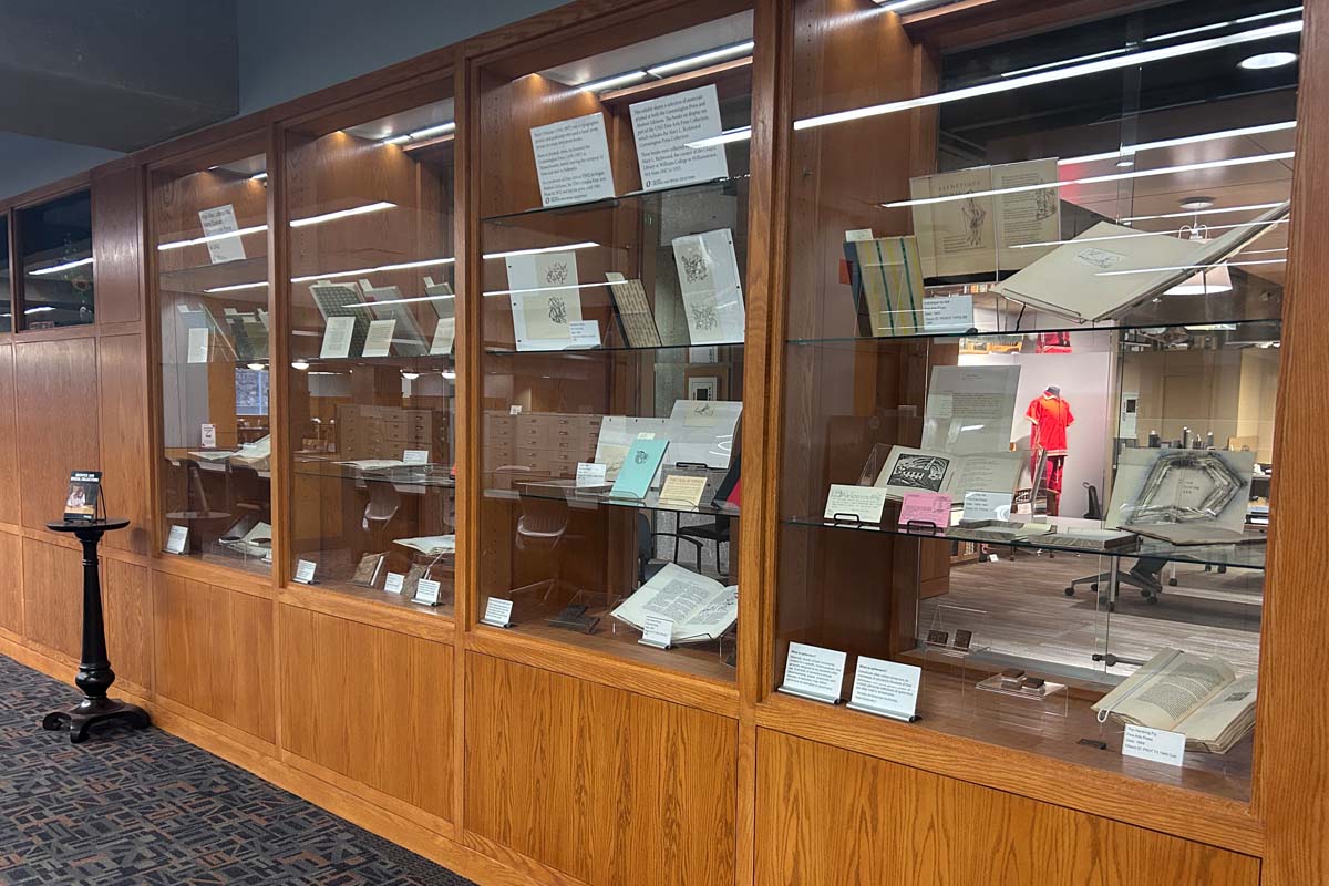 A library display case with historical documents and books on slanted shelves, each with labels.