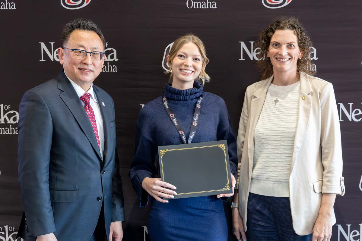 Three people stand in front of a University of Nebraska Omaha backdrop, with one holding a certificate.