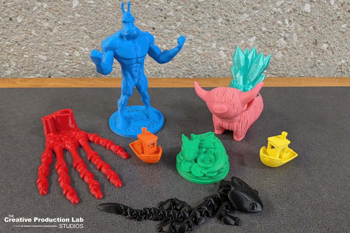 Colorful 3D printed models including a blue humanoid figure, a pink pig with crystals, boats, a dragon, a hand, and a scorpion on a gray surface.