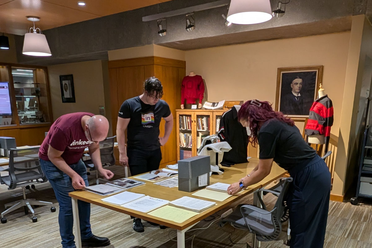 Three people examining documents laid out on a table in a library archive. Behind them are shelves filled with books and a framed portrait on the wall.