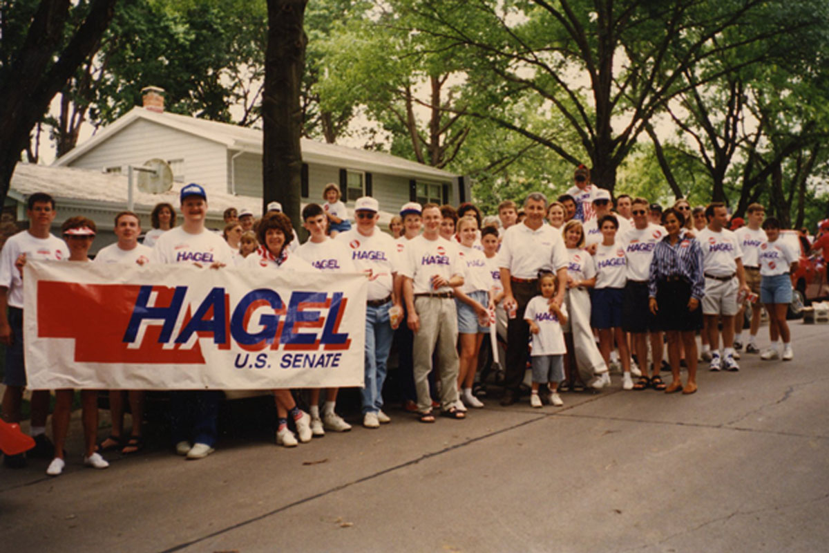 A group of people holding a parade banner for Chuck Hagel. They are standing on a city street and are looking at the camera and smiling.
