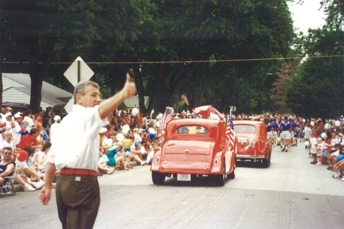 A man at a parade giving the crowd a thumbs up. He is wearing a white shirt and brown pants. Ahead of him there are small, red cars and on both sides of the street there are crowds.