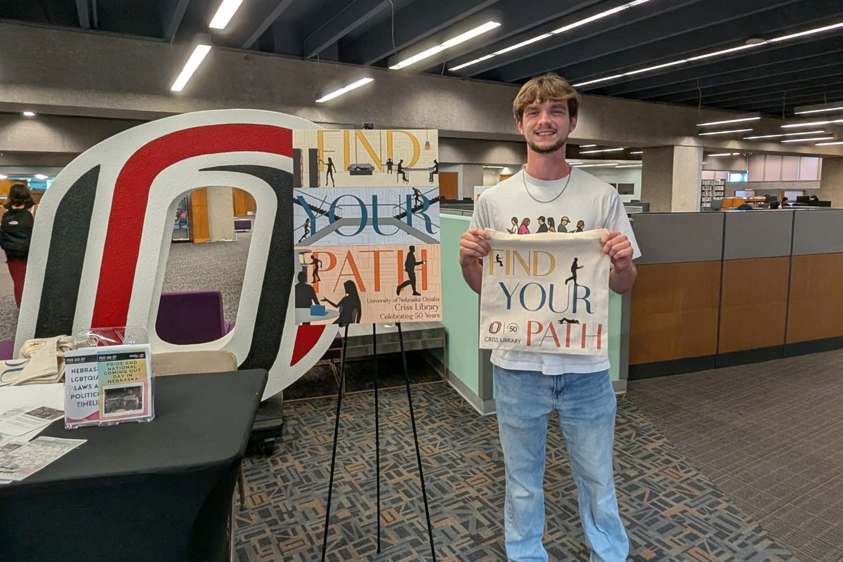 Young man holding a “Find Your Path” tote bag in a library beside a matching Criss Library anniversary poster. He has brown hair and is smiling at the camera.