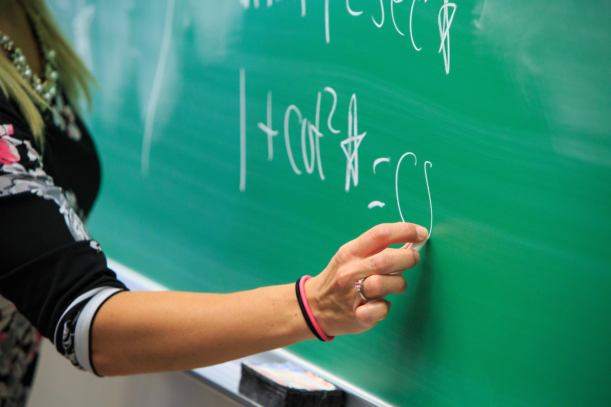 instructor's hand writing on green chalkboard with white chalk. mathematics formula in background on chalkboard.