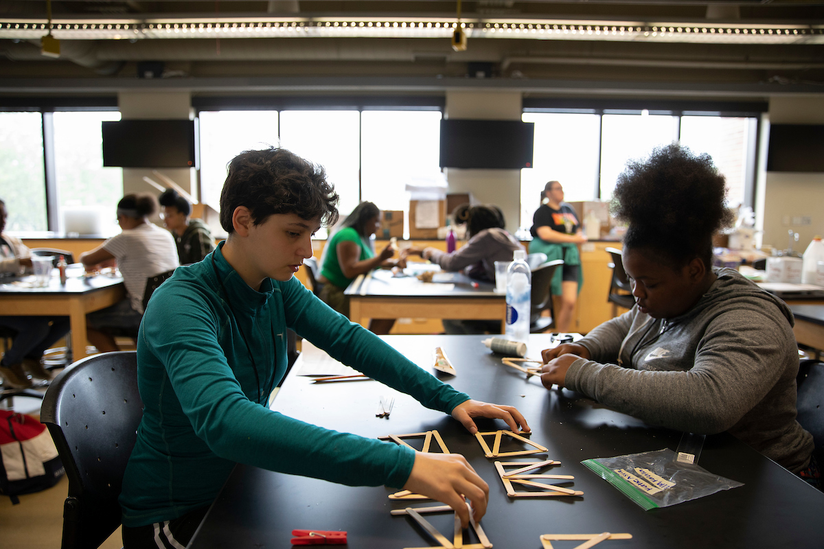 two students, sitting at a desk building a bridge out of popsicle sticks and glue.