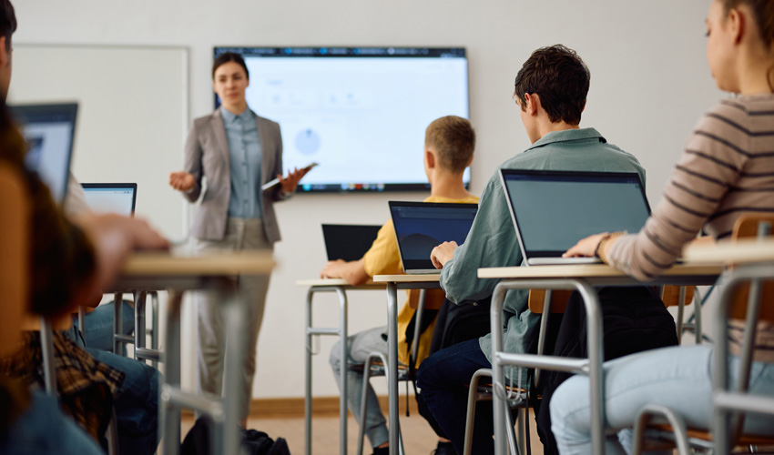 teacher standing at the front of a full classroom while students listen.