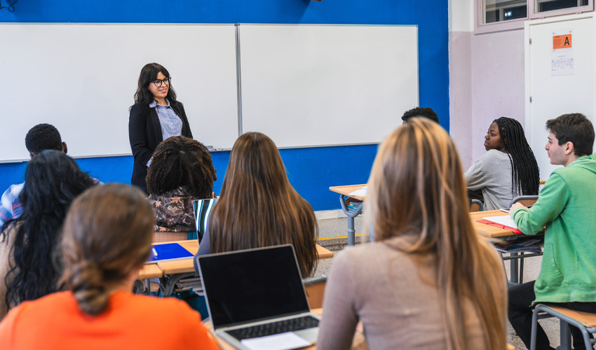 teacher standing at the front of a full classroom while students listen.