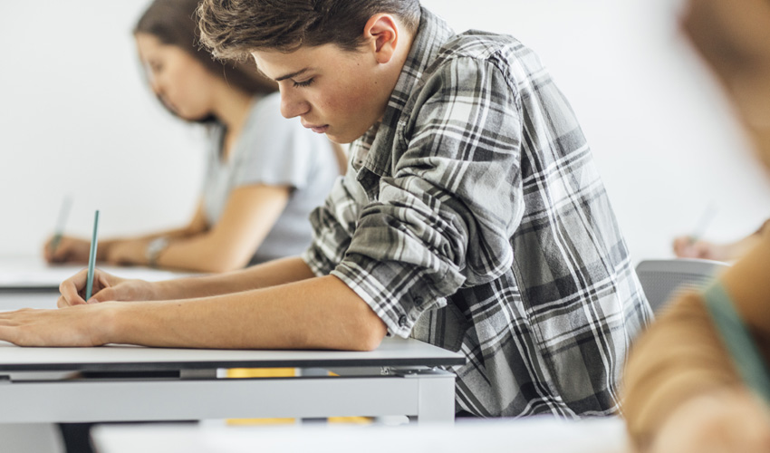 student focused intently on working on a paper, while seated at a desk in a classroom.
