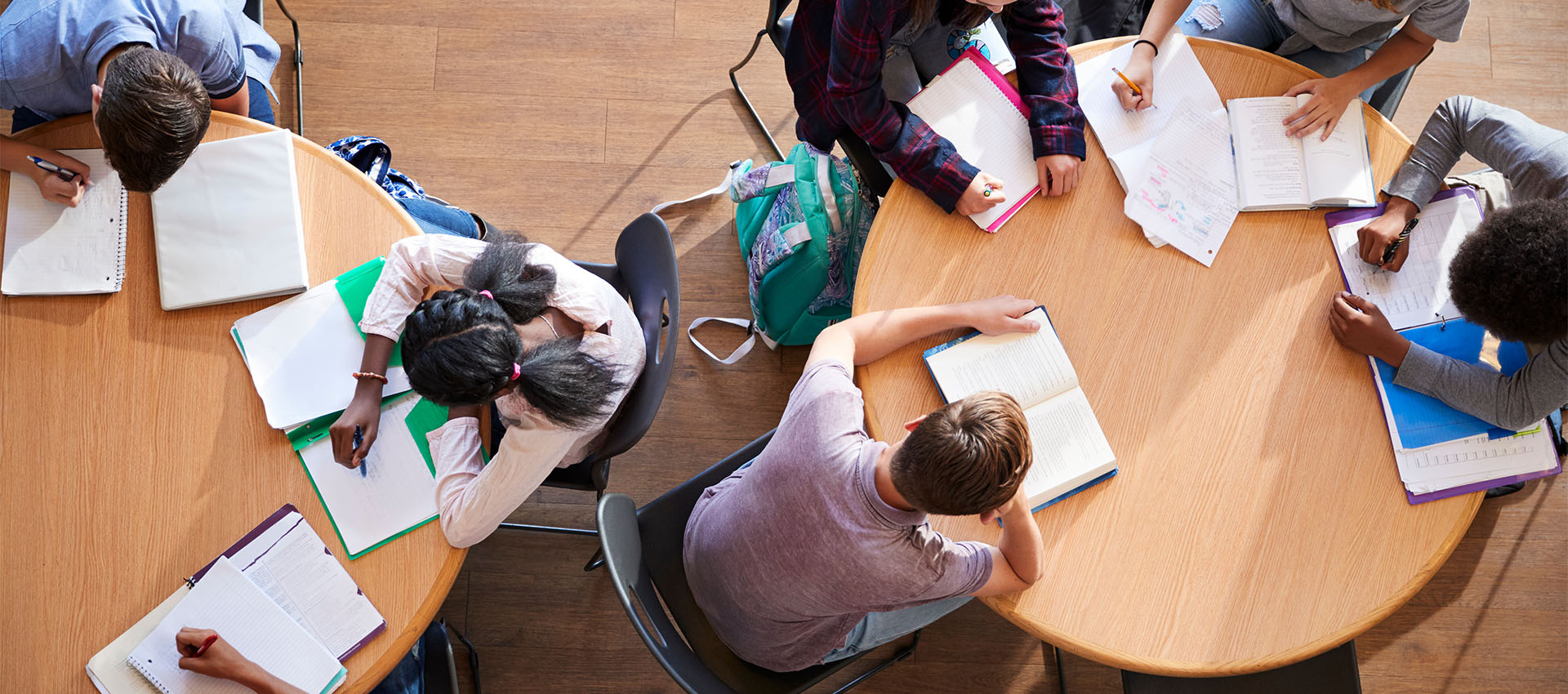 several students seated at 2 tables working on papers with open books, seen from above.