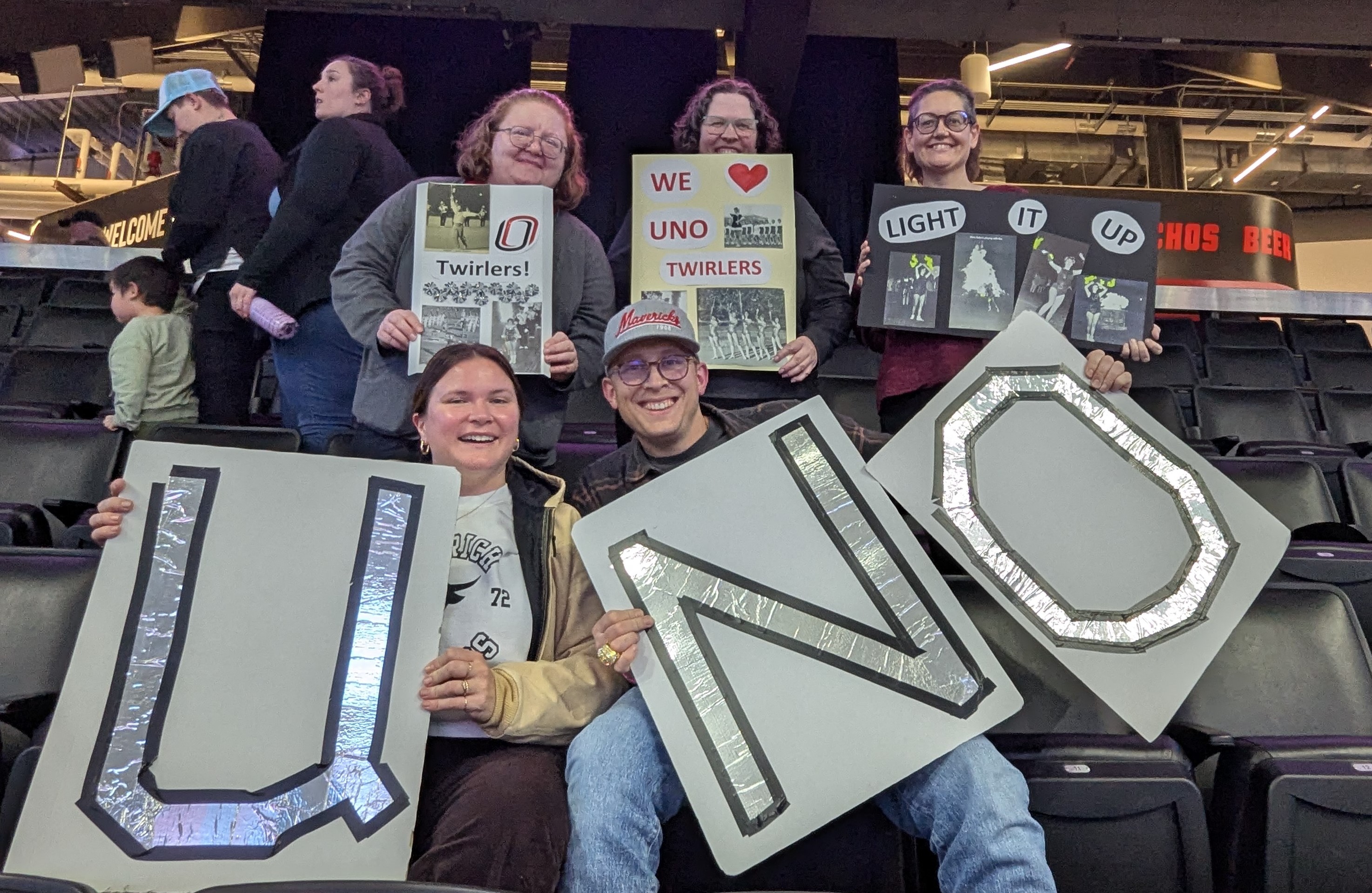 Six UNO basketball fans, standing in the bleachers of the Baxter Arena holding up signs in support of UNO basketball.
